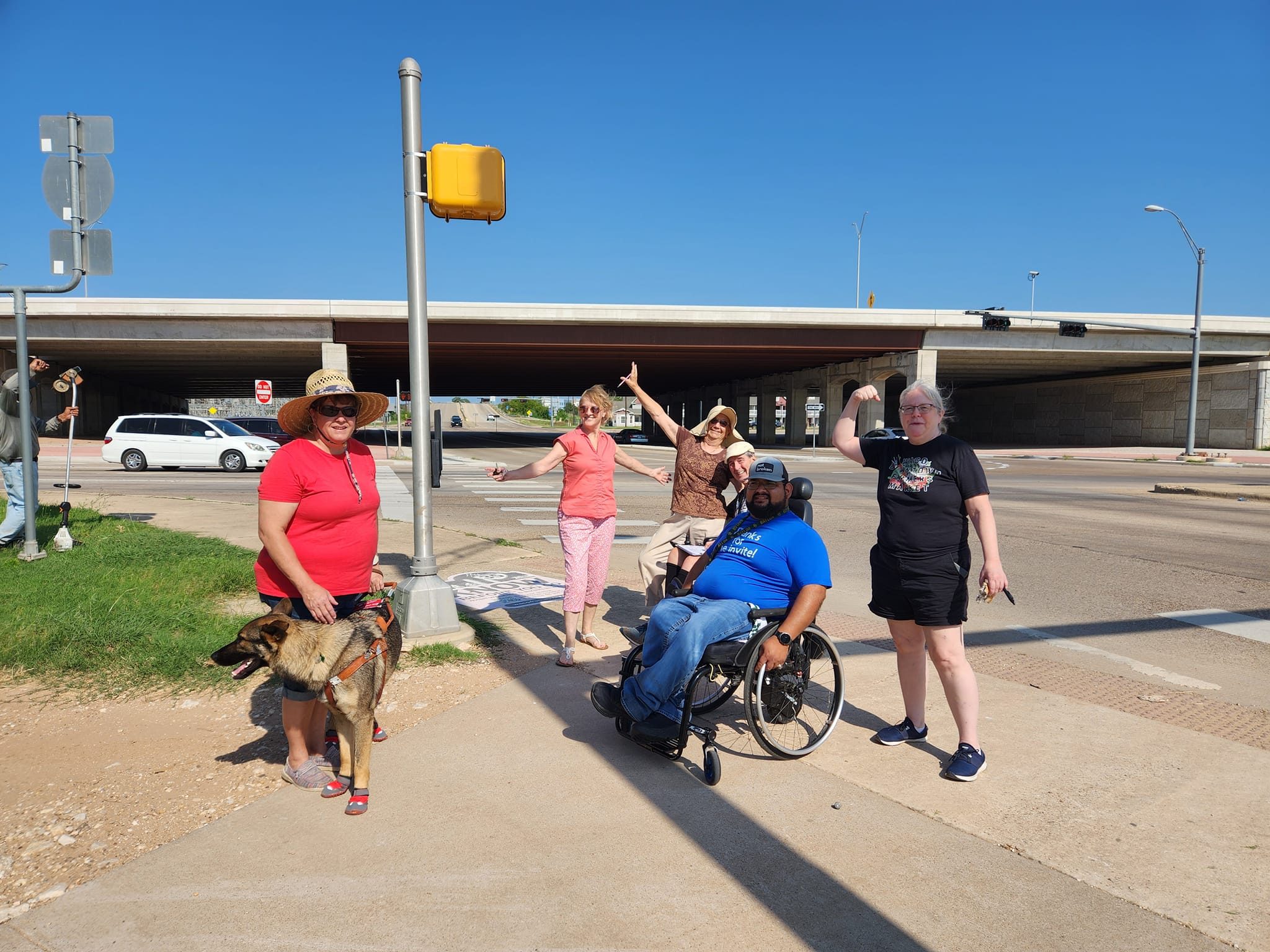 Four women standing, three in a celebratory pose, and two men in wheelchairs wearing hats and T-shirts, outdoors at an intersection near a highway overpass